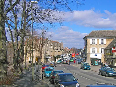 Witney High Street on a sunny day, with trees, shops and parked cars